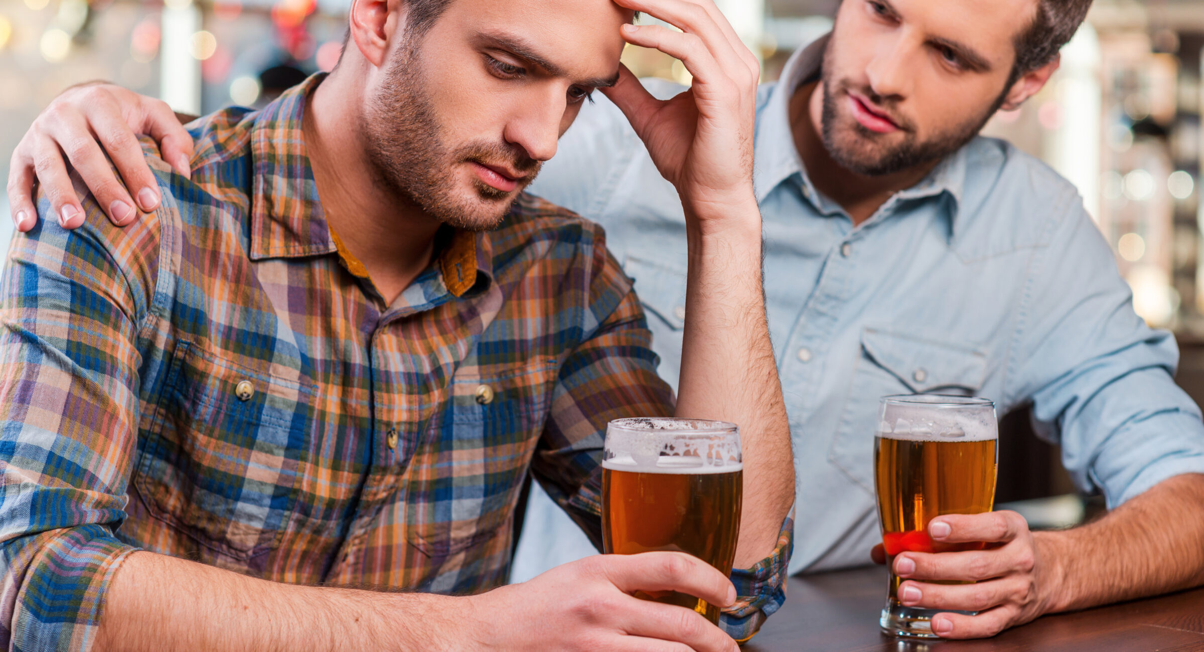 Depressed young man sitting at the bar counter and holding head in hand while being consoled by his friend sitting near him