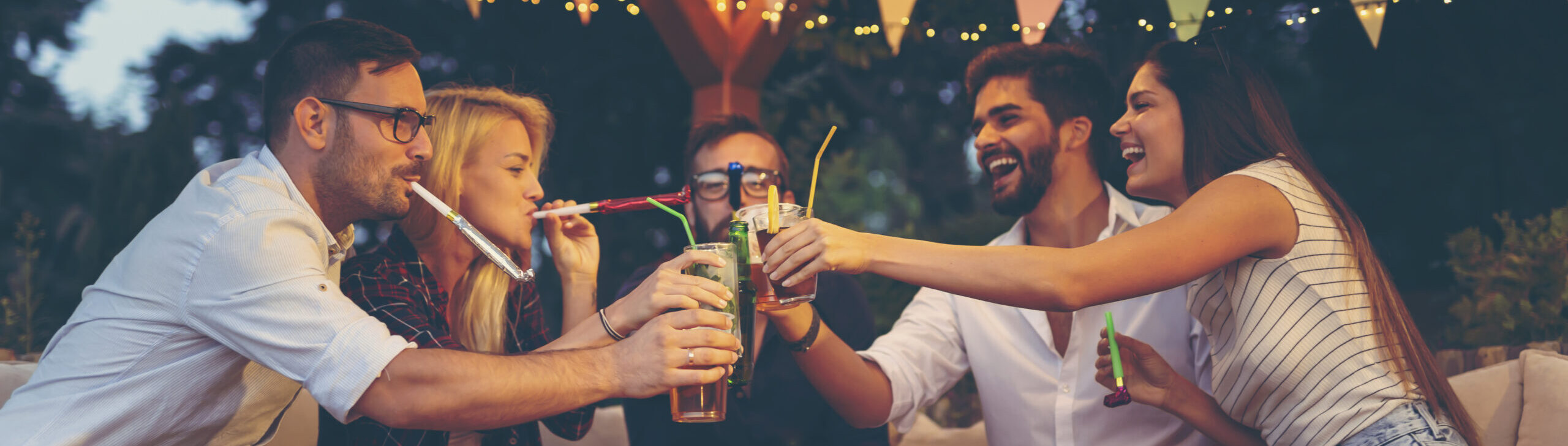 Group of young friends blowing party whistles, making a toast and having fun at an outdoor summertime party